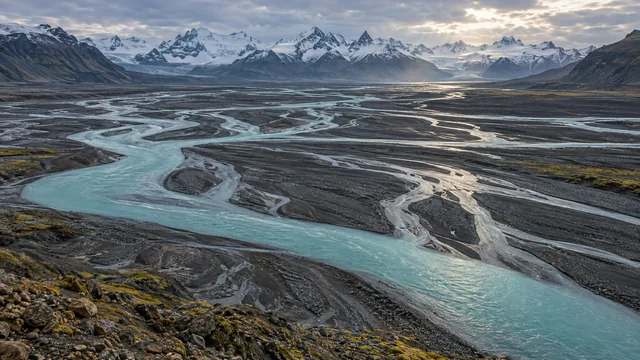 Turquoise glacial channels weave across a dark volcanic valley below distant snow-covered mountains